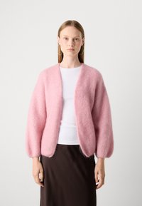 Cardigan fluffy di un rosa chiaro, realizzato in un materiale morbido con una vestibilità comoda, apertura frontale e maniche a palloncino, abbinato a una maglietta bianca e una gonna marrone.
