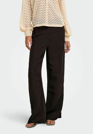 Personne portant un pantalon noir à jambes larges, un haut beige texturé à manches longues, des bagues aux doigts et des sandales beiges à bout ouvert.