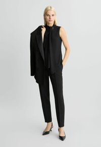 Zwart op maat gemaakt pak met een getailleerde mouwloze top met een gedrapeerde halslijn, slim-fit broek en puntige hakken.