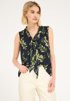 Femme portant un chemisier sans manches bleu marine à fleurs avec un nœud à l'avant et un pantalon blanc, debout avec une main dans la poche.