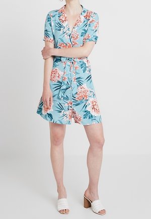 Kobieta ubrana w niebieską sukienkę z krótkim rękawem, zapinaną na guziki, z różowym kwiatowym wzorem i paskiem w talii, w połączeniu z białymi sandałami na obcasie.