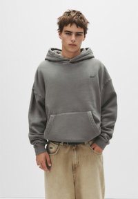 Grauer Hoodie aus Baumwollmischung mit einer Vordertasche und Kordelzugkapuze. Kombiniert mit beigen, lockeren Hosen, die ein dezentes Logo-Detail aufweisen.