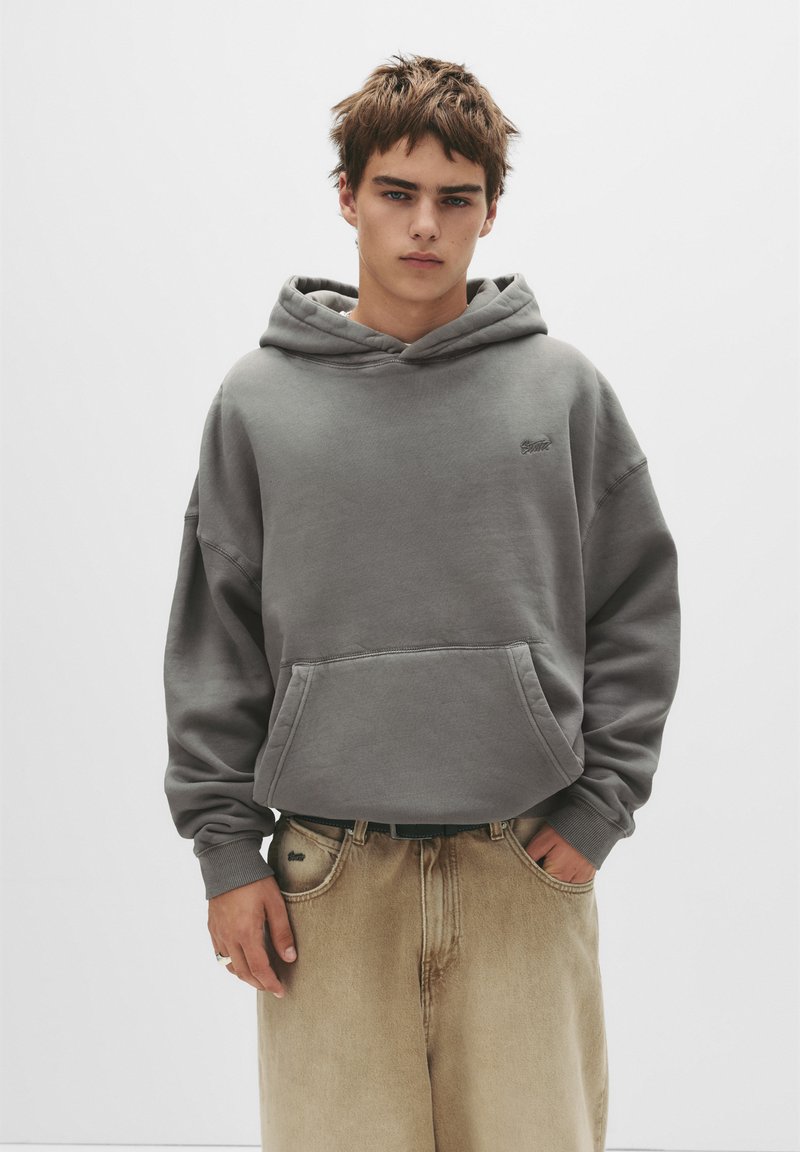 Grauer Hoodie aus Baumwollmischung mit einer Vordertasche und Kordelzugkapuze. Kombiniert mit beigen, lockeren Hosen, die ein dezentes Logo-Detail aufweisen.