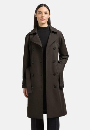 Kobieta w ciemnobrązowym, dwurzędowym płaszczu trencz z paskiem, ubrana na czarno, stojąca na tle jasnego, gładkiego tła.