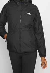 Chaqueta acolchada negra con cuello alto, cremallera frontal y cintura elástica. Presenta el icónico logo blanco de Adidas en el pecho izquierdo.