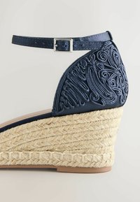 Sandale à talon compensé bleu marine avec semelle en toile tressée, dessus en tissu brodé et bride de cheville ajustable avec boucle argentée.