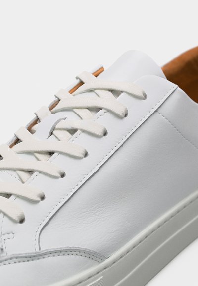 Μινιμαλιστικό λευκό δερμάτινο αθλητικό παπούτσι με λευκά κορδόνια, ταμπά εσωτερική επένδυση και διακριτικές ραφές σε λευκό φόντο.