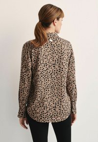 Koszula z długim rękawem wykonana z gładkiego materiału w odcieniu beżowym z czarnym, rozproszonym wzorem w kropki; posiada kołnierzyk oraz zapięcie na guziki z tyłu.