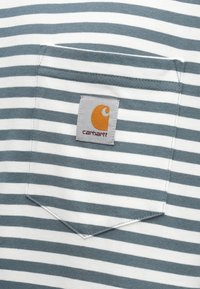 Ριγωτό γκρι και λευκό ύφασμα με πλακέ τσέπη που φέρει το λογότυπο Carhartt. Μαλακή υφή βαμβακιού με κανονικό, καθημερινό σχέδιο.