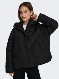 Woman wearing a black quilted jacket with snap buttons and zipper, adjusting her hair, standing against a plain light background.