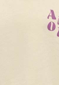 T-shirt in cotone beige con testo viola audace e design curvo, parzialmente oscurato. Il tessuto ha una texture liscia e una vestibilità casual.