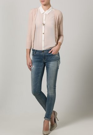 Cardigan à côtes rose clair sur une blouse blanche à col, assorti à un jean skinny bleu déchiré et des talons hauts beiges. Collier simple visible.