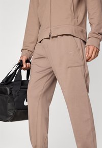 Φόρμα Nike σε μπεζ ύφασμα, με ζακέτα με φερμουάρ και παντελόνια με στενή γραμμή και διακριτική λεπτομέρεια λογότυπου. Περιλαμβάνει τσέπες και διακοσμητικές ραφές.