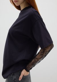 Schwarzes Baumwoll-T-Shirt mit weiten Ärmeln, das mit einem Spitzenüberzug an den Ärmeln versehen ist. Lockere Passform und runder Halsausschnitt.