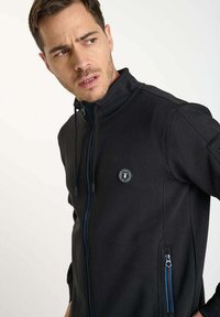 Veste noire à fermeture éclair devant avec col haut, accents bleus, poches latérales et patch logo. Tissu lisse avec un design ajusté.