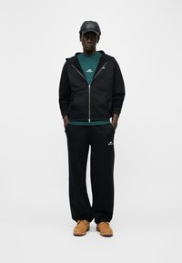 Νεαρός άνδρας φορώντας μαύρο φούτερ με κουκούλα, μαύρη φόρμα, πράσινο πουκάμισο, μαύρο καπέλο και καφέ εργοστασιακά παπούτσια, στέκεται μπροστά σε απλό ανοιχτόχρωμο φόντο.