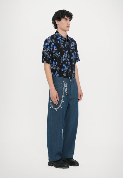 Kortärmad svart skjorta med blå blommönster, kombinerad med vida blå jeans som har en dekorativ kedjedetalj på fickan.