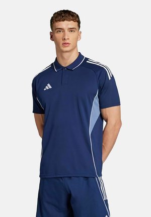 Giovane uomo che indossa una polo sportiva Adidas blu navy e pantaloncini, in piedi con le braccia dietro la schiena su sfondo bianco.