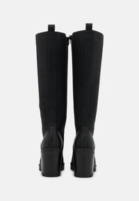 Bottes noires montantes jusqu'aux genoux avec une finition à texture lisse, une fermeture éclair et un talon épais. Présentent des coutures contrastantes le long des côtés.