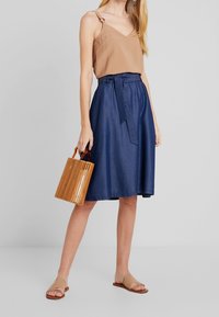 Nainen, jolla on yllään beigessä ilman hihoja oleva paita, sininen midi-hame vyöllä, kantaa puista ristikko käsilaukkua ja beigejä liukuvatsandaaleja harmaalla taustalla.