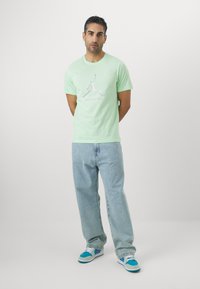 T-shirt in cotone verde chiaro con una stampa grafica sul davanti. Abbinato a jeans di denim azzurri e sneakers multicolori.