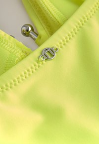 Jasně žlutá látka s hladkou texturou má dekorativní švy a stříbrný kovový doplněk uprostřed, což design vylepšuje.