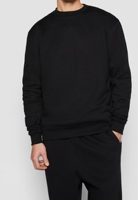 Sudadera negra de mezcla de algodón. Cuenta con cuello redondo, puños y dobladillo acanalados, ajuste relajado y textura suave. Sin patrones visibles.