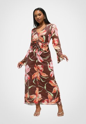 Frau trägt ein langärmeliges braunes Kleid mit großem pink-orangefarbenem Blumenmuster, Drehdetail an der Taille und dünnen Absatzsandalen.