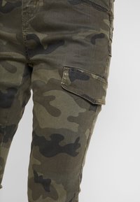 Pantalones cargo de camuflaje en verde oscuro y negro con una tela texturizada, con bolsillos laterales y un diseño ajustado.