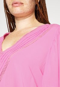 Blusa rosa com decote em V, apresentando pregas e detalhes em renda ao longo do decote. Tecido leve com uma textura suave.