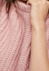 Grober Strickpullover aus weichem rosafarbenem Stoff mit hohem Kragen, der ein strukturiertes Muster und sichtbare Nähte zeigt, teilweise von einer Hand verdeckt.