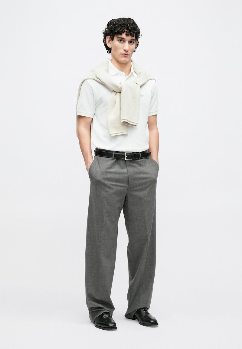 Weißes Poloshirt, graue, maßgeschneiderte Hose, beigefarbener Pullover lässig über die Schultern gelegt. Schwarzer Gürtel, schwarze Schuhe. Geradlinige Silhouette, minimale Details.