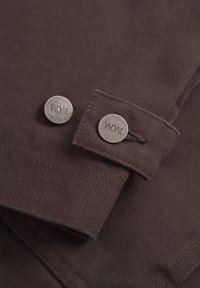 Rjava tkanina z teksturiranim finišem, ki vsebuje dva kovinska gumbka z vgraviranim "W.W.". Oblikovanje vključuje trak z gumbnim zapiranjem.