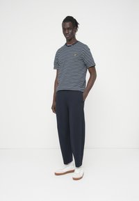 Κοντομάνικη μπλούζα ναυτικού στυλ με κίτρινο λογότυπο, σε συνδυασμό με σκούρο παντελόνι φαρδύ και λευκά αθλητικά παπούτσια. Απλό φόντο, το μοντέλο στέκεται άνετα.