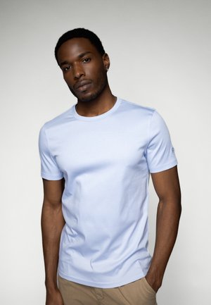 Helles blaues T-Shirt mit kurzen Ärmeln aus glattem Stoff, rundem Ausschnitt und dezenter Logodetails am Ärmel. Neutrale khakifarbene Hose sichtbar.