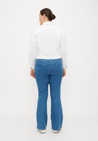 Chaqueta de denim blanca sobre jeans de campana azules. Los jeans cuentan con bolsillos traseros y un ajuste ligeramente relajado. Tejido texturizado, diseño limpio.