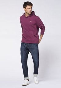Maroon hoodie with a kangaroo pocket and small logo, paired with dark jeans and white sneakers. The hoodie features a drawstring and ribbed cuffs.