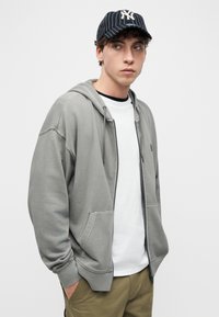 Νεαρός άνδρας που φοράει γκρι φούτερ με φερμουάρ, λευκό t-shirt, χακί παντελόνια και μαύρο και άσπρο ριγέ καπέλο μπέιζμπολ κοιτάζοντας προς τα πλάγια.