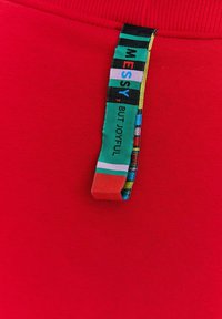 Czerwony materiał z bliska z wielokolorową, tkana metką z napisem „BAŁAGAN, ALE SZCZĘŚLIWY”, z różnorodnymi poziomymi wzorami w intensywnych kolorach.