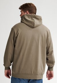 Felpa con cappuccio verde oliva, realizzata in tessuto morbido, caratterizzata da maniche lunghe, una cucitura posteriore e polsini e orlo a coste.