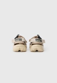 Sandalias exteriores beige con una suela de goma texturizada, correas ajustables y un acento negro en el talón con un diseño gráfico.
