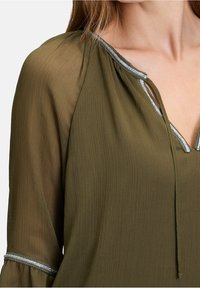 Blouse vert olive en tissu texturé, col en V et bordure argentée le long du col et des manches. Comprend un détail à cordon de serrage.