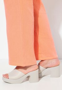 Beige blokhak mules met een brede band en open teen, gecombineerd met gestructureerde, koraalkleurige wijde broek. Gladde oppervlakte en stevig ontwerp.
