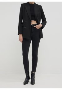 Femme portant un crop top noir, un blazer noir à double boutonnage, un jean skinny noir taille haute et des bottines noires, debout devant un fond blanc.