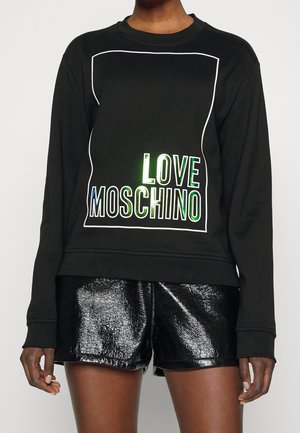 Persona vistiendo una sudadera negra de manga larga con el texto "LOVE MOSCHINO" dentro de un contorno rectangular blanco y pantalones cortos negros brillantes.