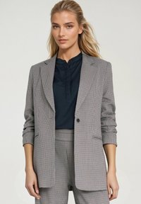 Szary blazer w kratę z wyraźnymi ramionami, klapami i zapięciem na jeden guzik, w połączeniu z granatową bluzką. Teksturowany materiał.