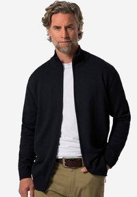 Námořnický kardigan na zip vyrobený z pleteného materiálu, s stojáčkovým límcem a žebrovanými manžetami. Nosený přes bílý tričko s khaki kalhotami.