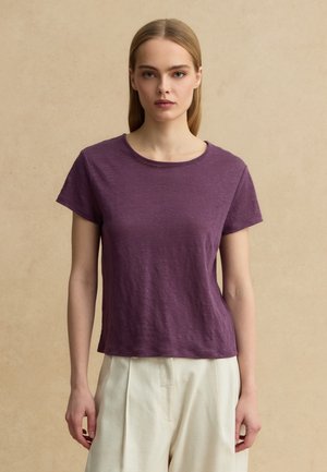 Joven mujer con cabello rubio lacio viste una camiseta morada lisa de manga corta y pantalones anchos de color crema, de pie frente a un fondo beige.