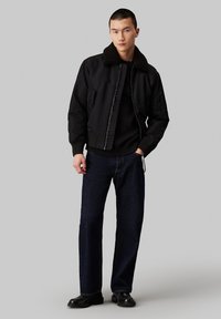 Chaqueta bomber negra con cuello de felpa, puños ajustados y frontal con cremallera. Llevada sobre un suéter negro y combinada con jeans oscuros y botas.