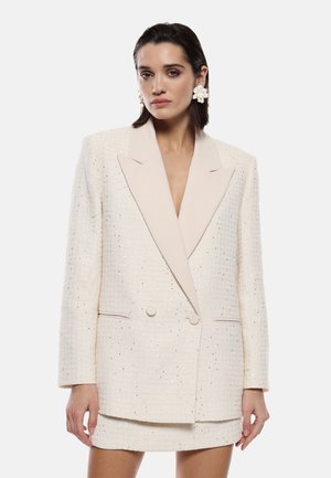 Blazer in tweed beige con un elegante risvolto chiaro, design doppiopetto e sottili accenti metallici, abbinato a una gonna corta coordinata.